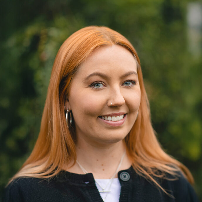 Portrait of Phoebe Wontner. Woman with red hair smiling at the camera. Wearing a black cardigan with a white top peaking through. Blurred background of greenery.