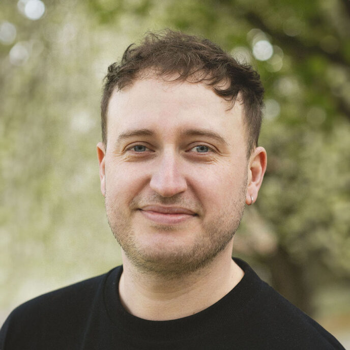 Portrait of man with short brown hair, wearing a black top. Background is blurred.
