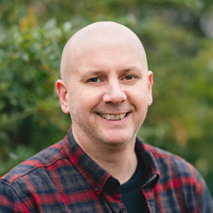 Portrait of a man wearing an open shirt with black undershirt, smiling at the camera. Background of blurred greenery.
