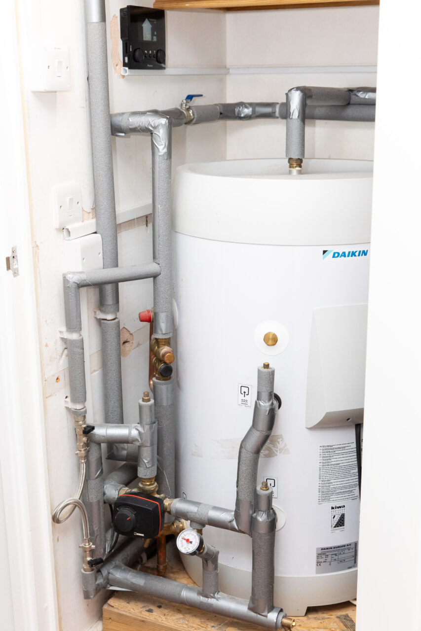 Photo showing a large hot water tank in an airing cupboard with insulated pipes and a control panel on the wall next to it.
