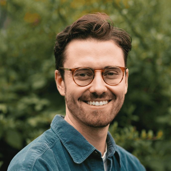 Portrait of man with brown hair and glasses smiling, wearing a teal overshirt with a green background