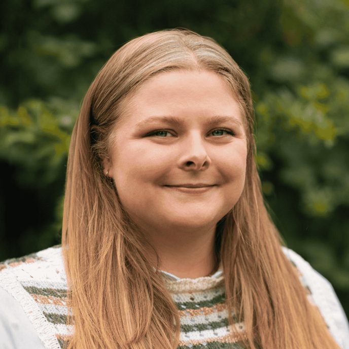Headshot portrait of woman with dark blonde hair smiling at the camera, wearing a white shirt with a knitted sleeveless vest. Behind her is a neutral green background of trees