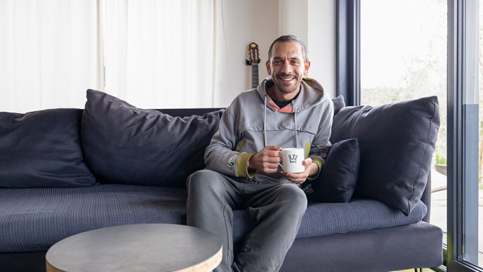 man smiling, holing a mug of tea, sat on a blue sofa in a clean and minimalist home.