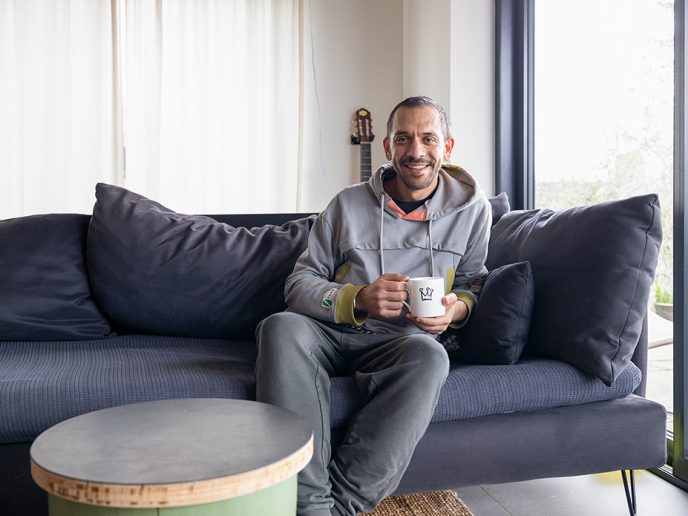 man smiling, holing a mug of tea, sat on a blue sofa in a clean and minimalist home.