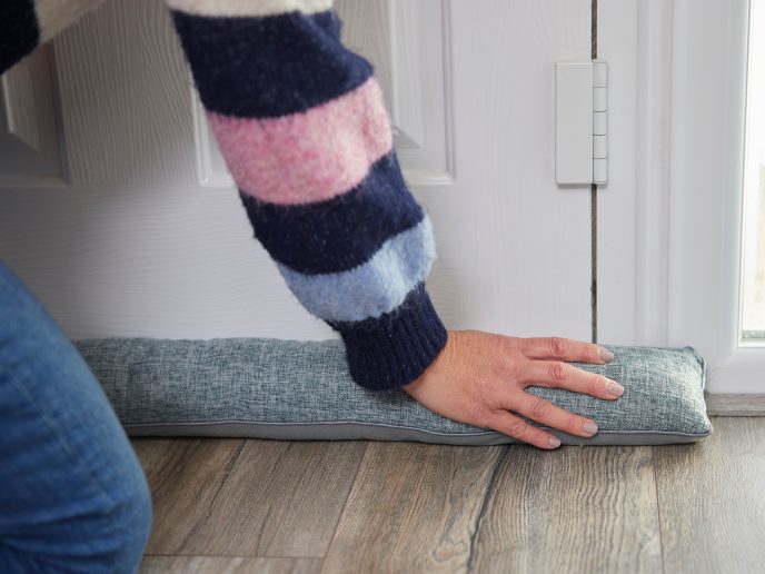person knelt in front of a closed door, pressing a material draught excluder to the base of the door to seal the gap between the door and floor.