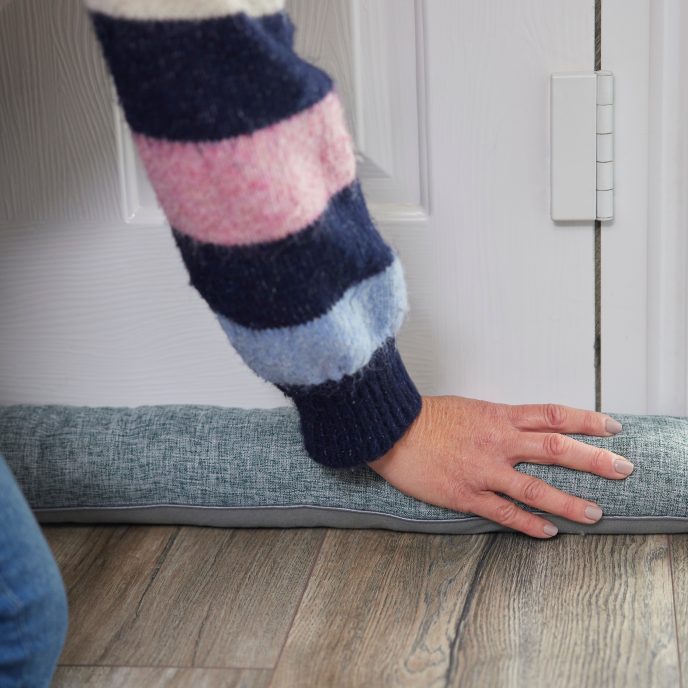 person knelt in front of a closed door, pressing a material draught excluder to the base of the door to seal the gap between the door and floor.