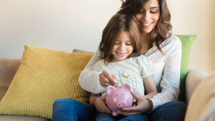 Woman and daughter sitting on a sofa, putting money into a piggy bank.