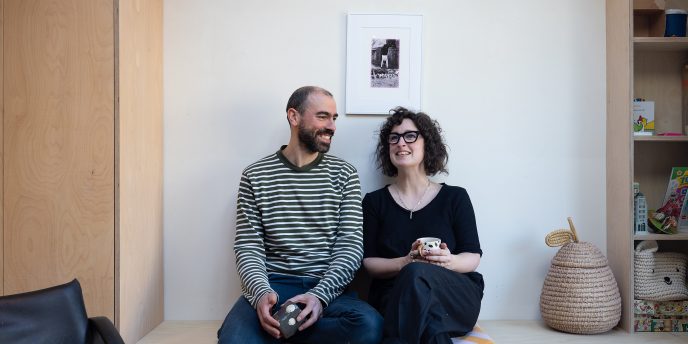 a laughing couple sat holding mugs on a minimalist wooden sideboard.