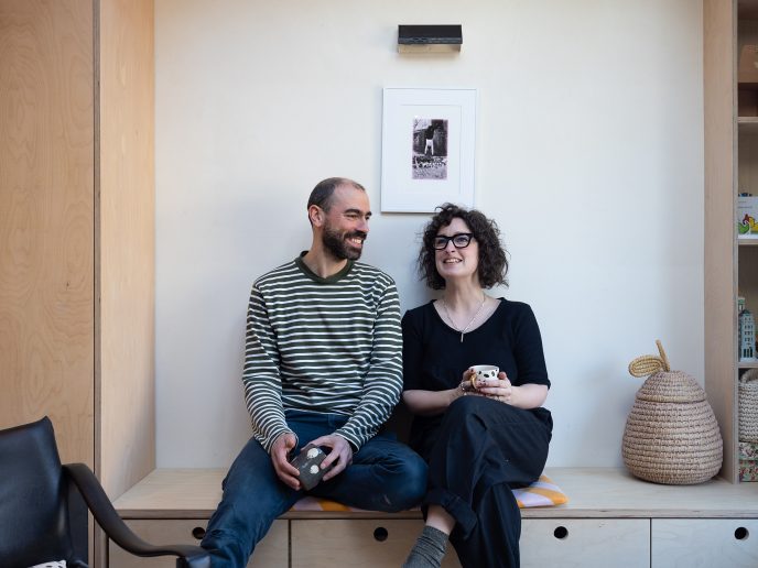 a laughing couple sat holding mugs on a minimalist wooden sideboard.