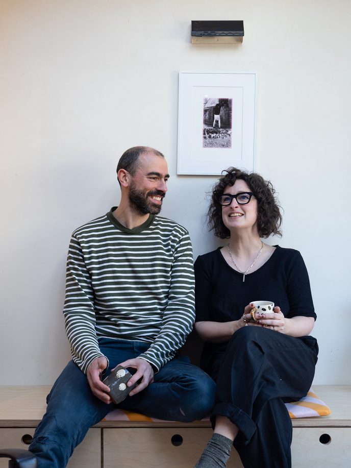 a laughing couple sat holding mugs on a minimalist wooden sideboard.