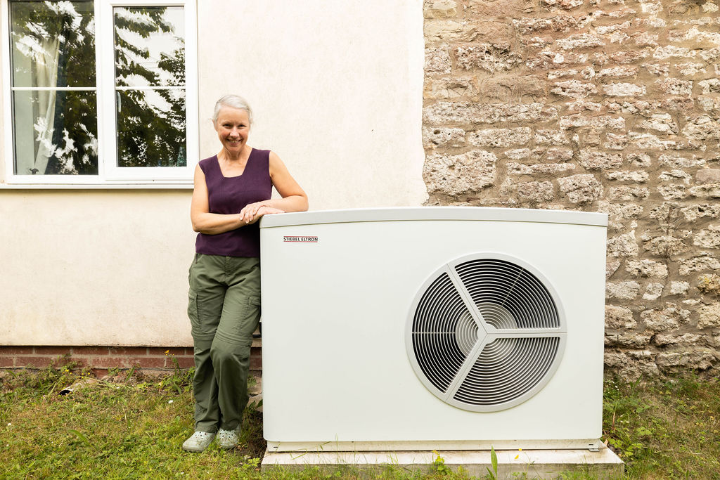 Neutrally toned photographer, of a woman standing next to her white heat pump. “Information about the Boiler Upgrade Scheme expansion for homeowners”
