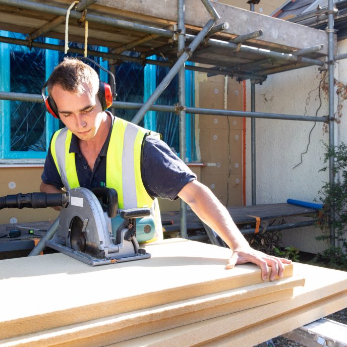 young man in hi-viz vest and ear defenders cutting EWI board outside with a table saw.