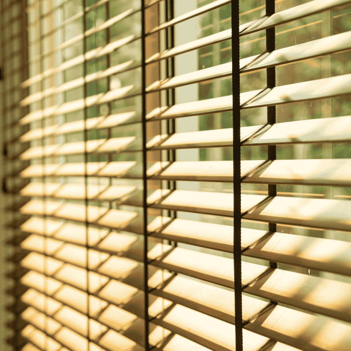 An upclose shot of a window with blinds, allowing sunshine to peak through.