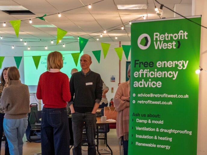 People networking at a community event. Retrofit West green standing banner in the forefront of the image, with green bunting on the ceiling.