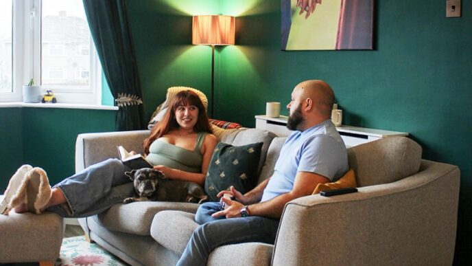 Bald, bearded man playing games console and brunette woman reading book, sat on grey sofa in green living room. Woman has feet up on a footstool and the pair are talking and smiling at each other.